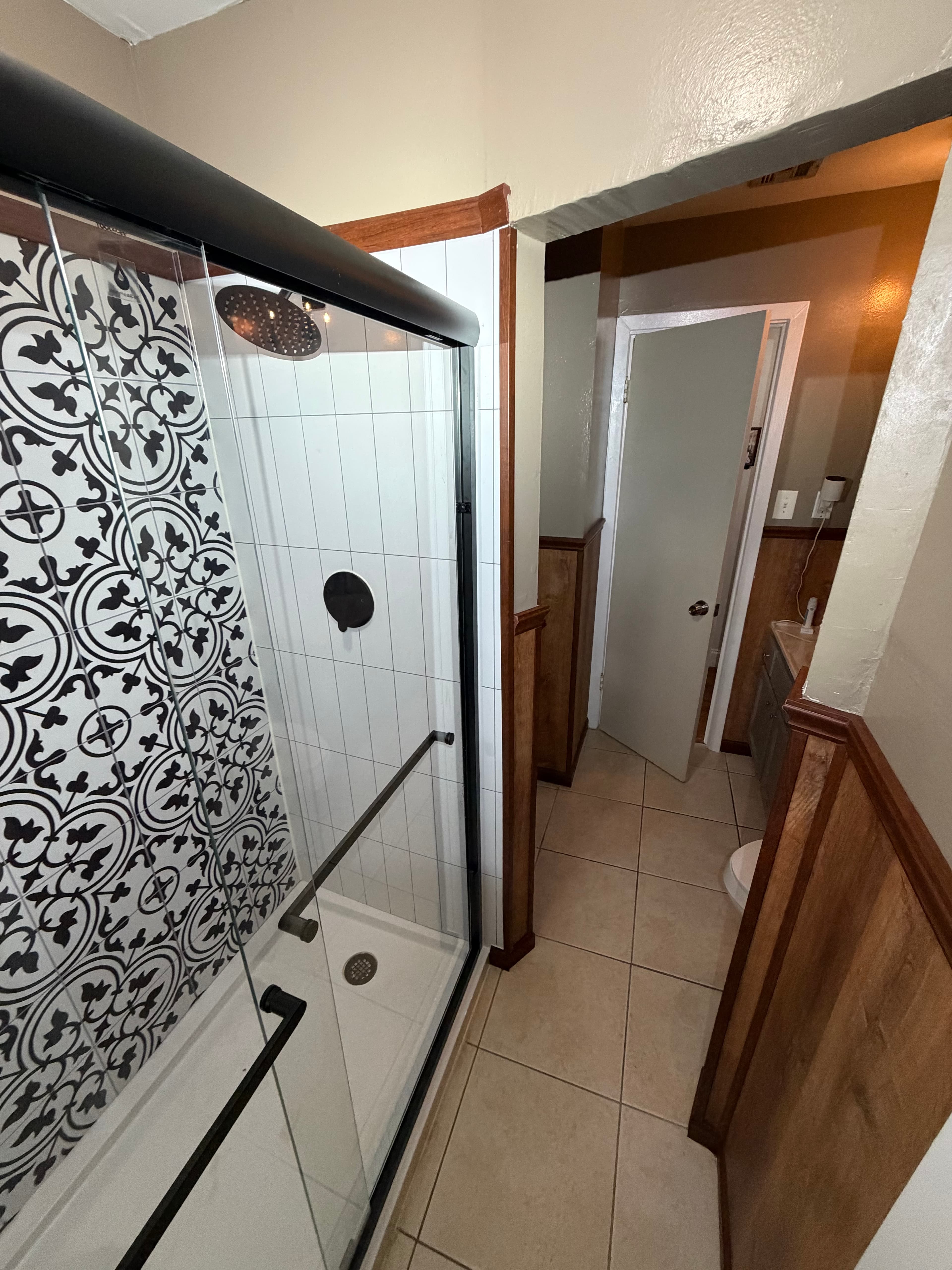 Modern shower featuring black and white patterned tiles beside a wood paneled bathroom hallway.