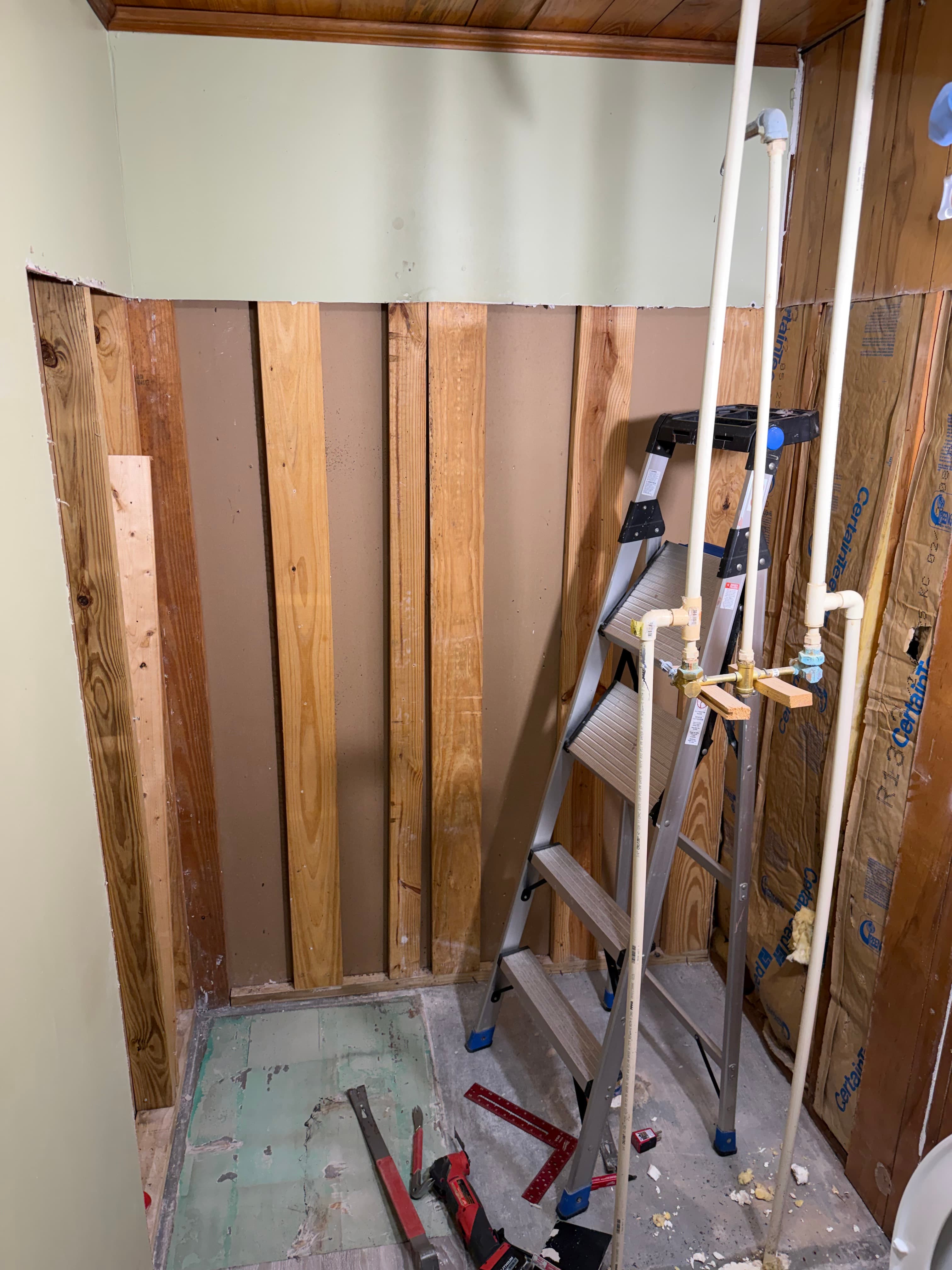 Interior room renovation featuring exposed wooden wall studs, vertical plumbing pipes, and a metal stepladder.